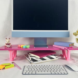 May include: A pink computer monitor stand with curved legs supports a blue and white computer monitor. The stand holds books, a small figurine, and colorful highlighters. A keyboard, mouse, and sticky notes are in front.