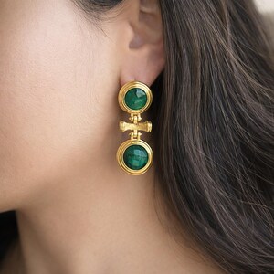 May include: Gold-toned earrings with a two-tiered design. Each tier features a round, faceted, emerald-green stone set within a gold frame. The earrings are connected by a decorative gold bar. Jewelry.