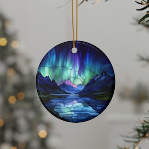 May include: Round ceramic ornament featuring a stained-glass style depiction of the Northern Lights over a mountain range and lake. The design showcases vibrant blues, greens, and purples. A gold string is attached for hanging. The ornament is displayed on a Christmas tree.