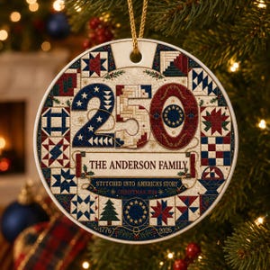 Op de afbeelding: Een ronde keramische kerstornament met een quilt-geïnspireerd patroon in rood, wit en blauw. Het getal 250 is prominent aanwezig, met de tekst THE ANDERSON FAMILY, STITCHED INTO AMERICA'S STORY en CHRISTMAS 2026 eronder gedrukt.