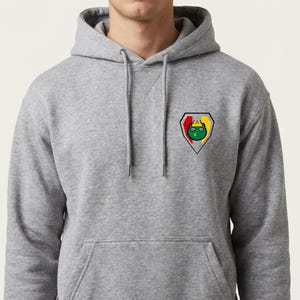 May include: A gray hooded sweatshirt with a front pocket and a drawstring. The sweatshirt features a small, colorful emblem on the chest. The emblem is a shield shape with a green cartoon character and yellow, red, and black accents.