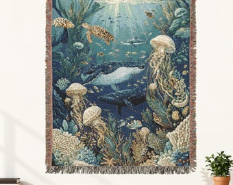 May include: A woven throw blanket featuring a detailed underwater scene with a whale, a sea turtle, a shark, and multiple jellyfish swimming among coral reefs and seaweed in shades of deep blue, teal, and cream with a fringed edge finish.