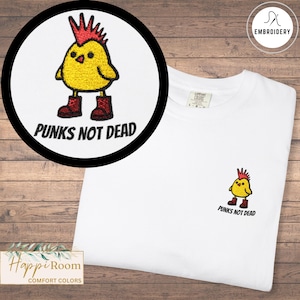 Può includere: Una t-shirt bianca con un ricamo raffigurante un pulcino giallo con cresta rossa e stivali rossi. Sotto il pulcino, il testo PUNKS NOT DEAD è scritto in lettere maiuscole nere. Il disegno è posizionato sulla parte sinistra del petto.