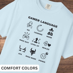 Op de afbeelding: Lichtblauw T-shirt met de tekst "GAMER LANGUAGE" en gaming-iconen. Het ontwerp bevat zinnen als "GG", "Loading", "Noob", "Rage Quit", "Level Up", "Healed", "AFK", "Boss Mode" en "Pwned". Het merk "COMFORT COLORS" is ook zichtbaar.