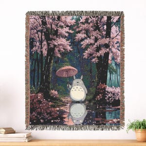 Può includere: Coperta tessuta raffigurante una scena del film d'animazione, Il mio vicino Totoro. Il design raffigura Totoro con un ombrello in una foresta con alberi di fiori di ciliegio rosa. La coperta ha un bordo sfrangiato ed è esposta su una superficie di legno.