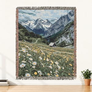May include: A woven throw blanket featuring a landscape scene of a mountain range with snow-capped peaks, a green valley, a small cabin, and a foreground filled with white daisies and yellow wildflowers. The blanket has a decorative fringed edge.