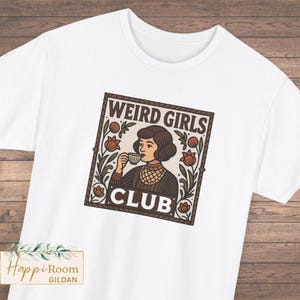 May include: A white short-sleeve t-shirt featuring a square graphic print of a person with short brown hair drinking from a small cup. The design includes floral elements and the text WEIRD GIRLS CLUB in a dark brown, cream, and green color palette.