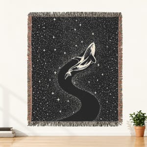 May include: A woven throw blanket featuring a black background with a white stippled design of a whale swimming through a starry night sky. The blanket has a fringed edge on all four sides and a textured, tapestry-like appearance throughout.