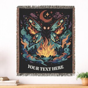 May include: Black throw blanket with a colorful illustration of a moth with flames, a city skyline, and the text "YOUR TEXT HERE". The moth has a detailed design with orange, blue, and yellow accents. The blanket has a fringed edge.
