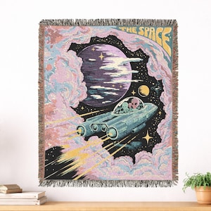 May include: A woven throw blanket featuring a space scene with a blue spaceship carrying two aliens, a large purple planet, and yellow stars. The text THE SPACE is printed in yellow at the top, surrounded by pink and blue clouds on a black background.