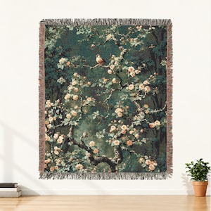 May include: A woven throw blanket featuring a dark forest green background with a detailed pattern of light peach-colored roses and small birds perched on winding branches. The blanket has a textured fringe along all four edges.