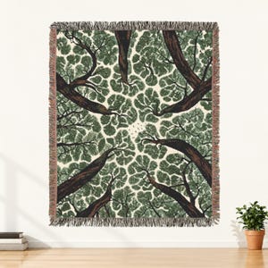 May include: A woven throw blanket featuring a low-angle perspective of tree trunks and branches reaching toward a cream-colored sky. The design displays dark brown bark and dense green foliage with a fringed edge on all four sides of the textile.