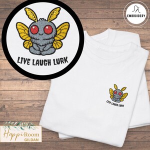 May include: A white t-shirt featuring an embroidered design of a grey moth with large red eyes and yellow wings. The moth is sitting in a meditative pose. Below the moth, the text reads "LIVE LAUGH LURK" in a bold, black, sans-serif font.