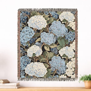 May include: A rectangular woven throw blanket featuring a dense floral pattern of hydrangea clusters in shades of light blue, white, and dark blue against a black background. The blanket has a textured weave and frayed edges on all four sides.