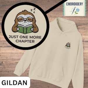 May include: Cream-colored hoodie with a front pocket and drawstring hood. The design features a cartoon sloth wearing glasses and reading a book, with the text "JUST ONE MORE CHAPTER". The same design is embroidered on the upper left chest.