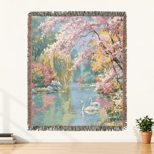 May include: A woven throw blanket featuring a landscape scene of two white swans swimming on a calm pond. The scene includes lush trees with pink and yellow blossoms, green foliage, and a soft blue sky, all finished with a fringed edge border.