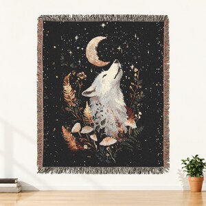May include: A woven throw blanket featuring a black background with a white wolf howling at a crescent moon. The design includes detailed illustrations of ferns, mushrooms, and small flowers in earthy tones, finished with a fringed edge.