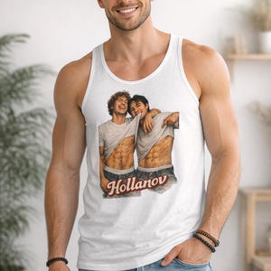 May include: White tank top featuring a graphic of two men with exposed abs, embracing. The word "Hollanov" is printed in a red, stylized font. The tank top is worn by a person in blue jeans.