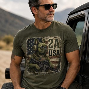 Peut inclure: Un t-shirt vert olive avec un motif représentant un bouledogue français portant un équipement tactique et un casque, tenant un fusil devant un drapeau américain vieilli. Le texte 2A USA et GREEN BULLDOG SHIRT CO est imprimé sur le devant.