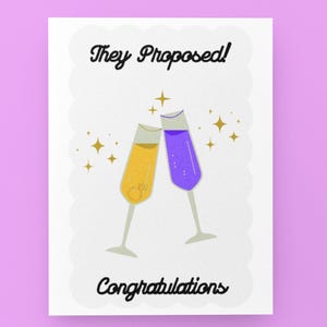 May include: A white greeting card with a scalloped edge displays the text "They Proposed!" and "Congratulations" in black script. Two champagne flutes are clinking, one filled with yellow liquid and the other with purple. A ring is visible in the yellow flute.