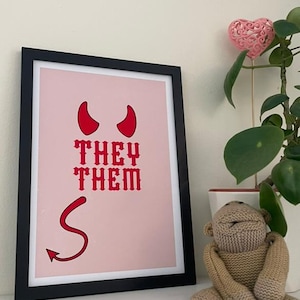 May include: A framed pink art print featuring red devil horns, a tail, and the words "THEY THEM" in red. The artwork is in a black frame, displayed on a white shelf, with a knitted monkey toy and a potted plant.