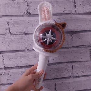 Puede incluir: Un palo de luz blanco y rojo con un diseño de estrella, cubierto con una funda de orejas de conejo de ganchillo hecha a mano. El mango tiene el texto "Stray Kids" impreso. El palo de luz se sostiene contra una pared de ladrillos.