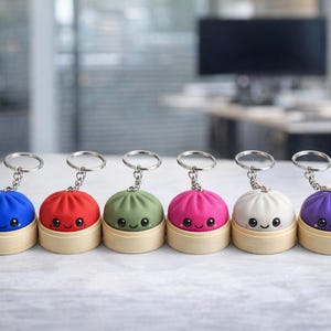 May include: Six round keychains featuring colorful dumpling-shaped charms with smiling faces. Each charm sits in a small, light-colored wooden steamer basket base and includes a silver-toned metal keyring attached by a short chain.