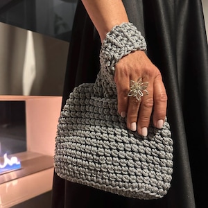 Puede incluir: Un bolso de mano gris tejido a ganchillo con un brazalete a juego. El bolso tiene un aspecto texturizado y tejido y un asa anudada. Un anillo de flores plateado se lleva en la mano. El fondo es un vestido negro.