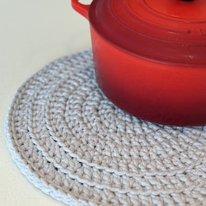 Op de afbeelding: Een grijze gehaakte pot onderzetter met een rode pot erop. De pot onderzetter is rond en heeft een textuur.