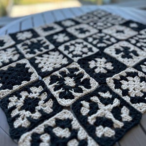 May include: A handmade crochet blanket featuring a repeating pattern of black and cream-colored squares. Each square showcases a floral design, creating a cozy and visually appealing textile. The blanket is perfect for adding a touch of handmade charm to any space.