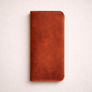 Puede incluir: Una funda rectangular de cuero marrón con un acabado envejecido. La funda está cosida alrededor de los bordes y tiene una esquina superior redondeada. El cuero presenta variaciones de color y textura.
