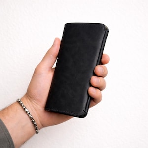 May include: A black leather wallet with a zip closure. The wallet is held in a hand, and a silver bracelet is visible on the wrist. The background is plain white.