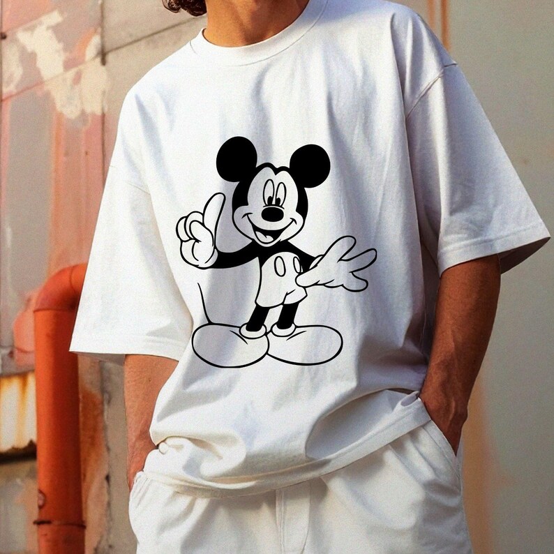 May include: White t-shirt featuring a black and white graphic of Mickey Mouse. The cartoon character is depicted with his signature ears, gloves, and shoes, giving a thumbs-up gesture. The shirt is a casual style.