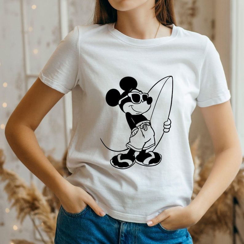 May include: White t-shirt featuring a black and white graphic of Mickey Mouse wearing sunglasses and holding a surfboard. The cartoon character is dressed in shorts and sandals. The shirt is a casual style.