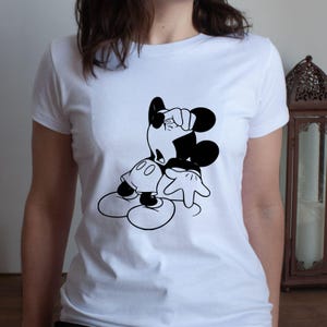 May include: White t-shirt featuring a black and white graphic of Mickey Mouse in a classic pose. The cartoon character is leaning with one hand on his head and the other hand extended. The shirt is a crew neck style.