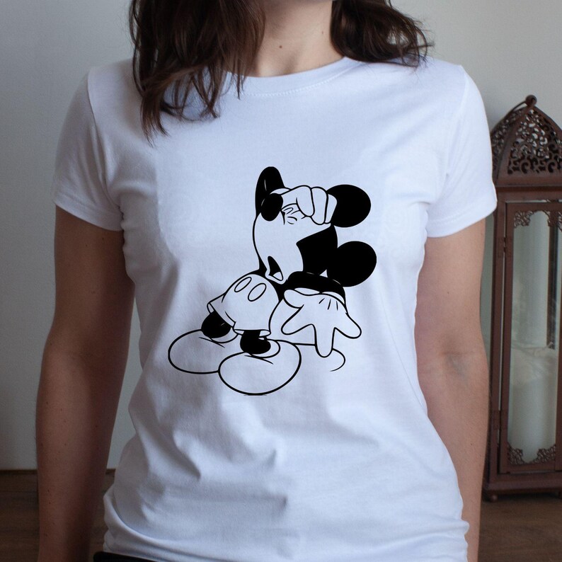 May include: White t-shirt featuring a black and white graphic of Mickey Mouse in a classic pose. The cartoon character is leaning with one hand on his head and the other hand extended. The shirt is a crew neck style.
