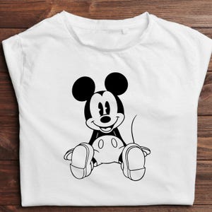 May include: White t-shirt featuring a black and white illustration of Mickey Mouse sitting with his legs crossed. The cartoon character has large black ears, a smiling face, and white gloves and shoes. The shirt is made of a soft material.