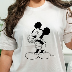May include: White t-shirt featuring a black and white graphic of Mickey Mouse holding a lollipop. The cartoon character is smiling and wearing a black outfit with white gloves and shoes. The shirt is a classic, casual style.