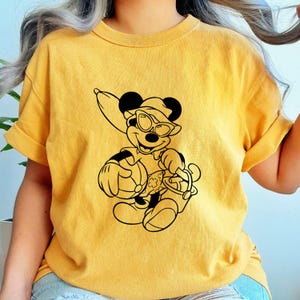 May include: Mustard yellow t-shirt featuring a black graphic of a cartoon mouse wearing sunglasses, a hat, and holding a beach bag. The mouse is smiling. The shirt has short sleeves and a crew neck.