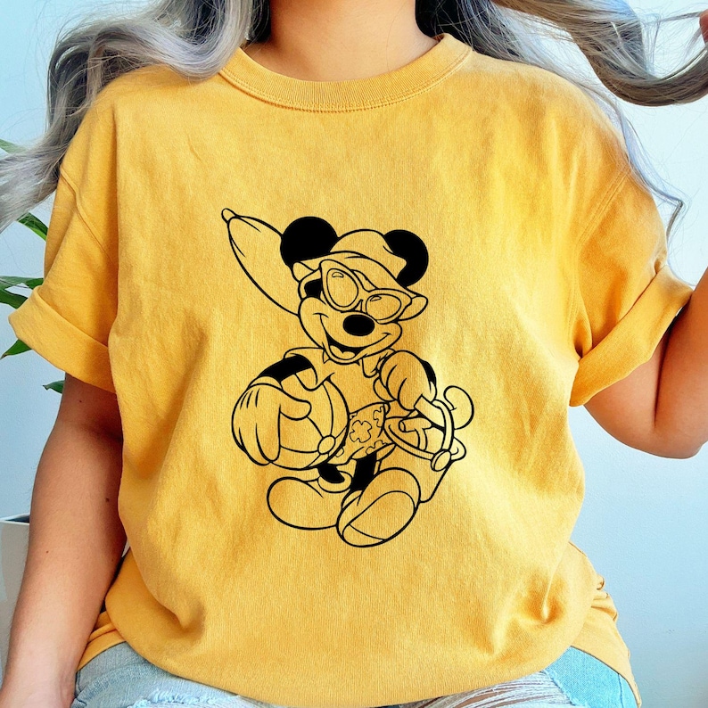 May include: Mustard yellow t-shirt featuring a black graphic of a cartoon mouse wearing sunglasses, a hat, and holding a beach bag. The mouse is smiling. The shirt has short sleeves and a crew neck.