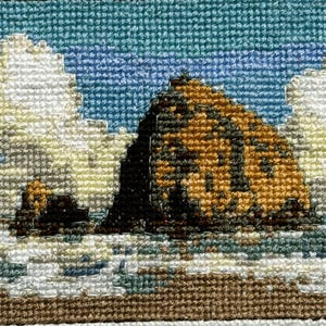 May include: Cross-stitch artwork depicting Haystack Rock, a large rock formation in the ocean. The scene includes a blue sky, white clouds, and waves. The rock is brown and black. The text "HAYSTACK ROCK" is stitched below the image.