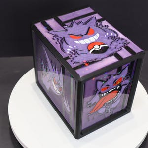 Puede incluir: Una caja negra y morada con Gengar de Pokémon. La caja tiene forma rectangular con frente y laterales transparentes, mostrando ilustraciones de Gengar. La parte superior de la caja tiene un diseño a rayas con un gráfico de Gengar y una Pokéball.