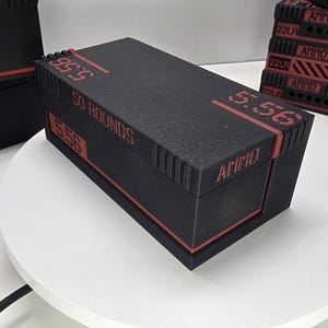 May include: Black rectangular container with red detailing and the word "AMMO" printed on the side. The container has the numbers "5.56" and "50 ROUNDS" in red. Other similar containers are in the background.