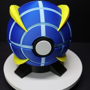 May include: A blue and yellow spherical object with a white and black center, resting on a white circular base. The sphere has light blue stripes and yellow accents. The base is supported by black, angular legs.
