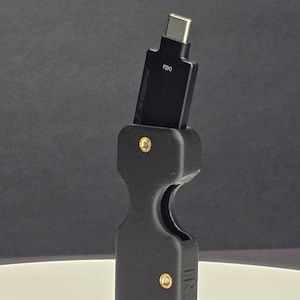 May include: Black USB-C adapter with the text "FIDO" and a key ring. The adapter is held in a black holder with gold-colored rivets and the text "IR-3D".