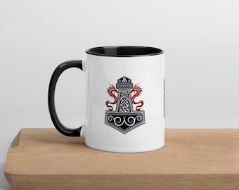 Puede incluir: Taza de cerámica blanca con interior y asa negros. La taza presenta un diseño detallado de un martillo con dragones rojos y intrincados nudos. El diseño está centrado en la taza, creando un efecto visual llamativo.