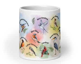 May include: White ceramic mug featuring watercolor illustrations of various North American birds, including a bluebird, cardinal, and American robin. Each bird is labeled with its name, set against a colorful, abstract background.