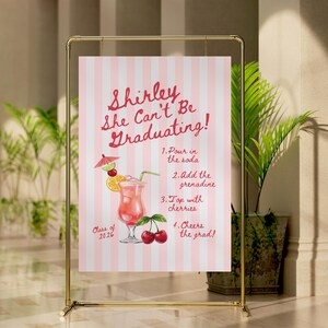 May include: A vertical rectangular sign with a light pink and white striped background. The text reads Shirley She Can't Be Graduating! followed by a numbered recipe for a cocktail with an illustration of a drink, cherries, and Class of 2026.