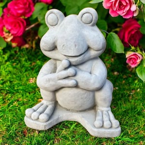 May include: A gray frog statue with its hands clasped, sitting on green grass. The frog has large, round eyes and a smiling expression. Pink roses are in the background.