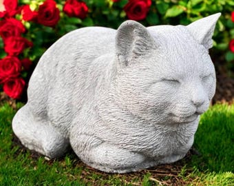 May include: A gray cat statue resting on green grass, with closed eyes and a relaxed posture. The cat sculpture is surrounded by red roses in full bloom, creating a garden scene. The statue is made of a textured material.